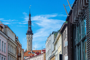 The iconic Tallinn Town Hall spire rises elegantly above the city, framed by colorful historic houses and modern architecture under a bright blue summer sky.