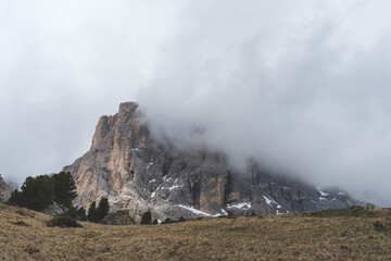 Dramatic view of Sassolungo mountain partly covered by clouds, with rocky cliffs and grassy alpine meadow in the Dolomites, Italy.