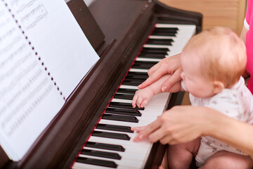 Young mother with light hair guides baby girls hands on piano keys. Casual clothes, soft light,...