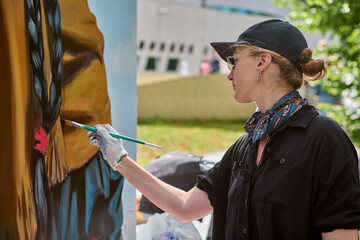 Young adult caucasian female artist in black shirt and cap painting mural outdoors. Woman focused,...