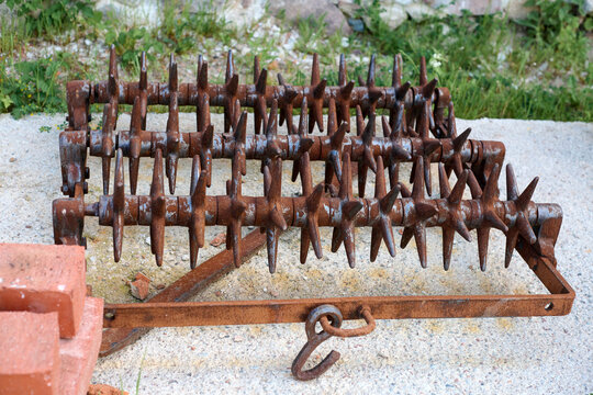 Close up of rusty spiked harrow agricultural farm implement used for surface tillage. Harrow resting on concrete surface, with greenery in background