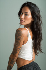 Fototapeta premium Woman with long dark hair and tattoos poses confidently in a light green setting while wearing a cropped white tank top and black shorts during daytime