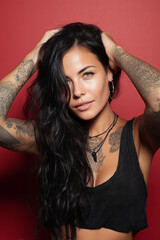 Young woman with long black hair and tattoos poses against a vibrant red background, showcasing her unique style and confidence