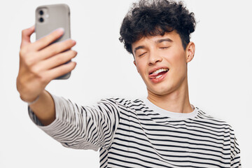 young man taking selfie with smartphone, wearing striped shirt, playful expression, white...
