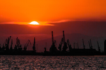 Sunset behind industrial port cranes in silhouette with glowing orange sky and sea reflections,...