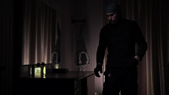 A man in black clothing appears and begins rummaging through a dresser drawer
