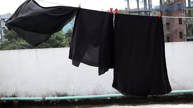 Black burqa hanging on clothesline, swaying in wind. Muslim woman's attire sun drying outdoors.