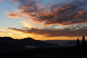 Fototapeta premium Breathtaking sunset paints sky with vibrant hues of pink, orange, purple, blending into blue morning sky. Silhouetted mountains, pine trees create serene backdrop. Tranquil and picturesque landscape.