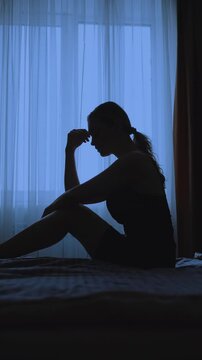 black silhouette upset unhappy woman sitting on bed dark room mood depression sadness emotion. Sad lonely Girl feels bad after party, insomnia migraine suffers headache. window curtains vertical video