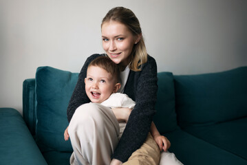 Smiling young mother hugging her cheerful son while sitting on a modern teal sofa at home, capturing a moment of joy and family love.