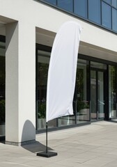 A mockup of a tall, blank white feather flag standing outside a modern storefront on a sunny day. The flag is gently waving in the breeze. The background shows a clean, modern building facade