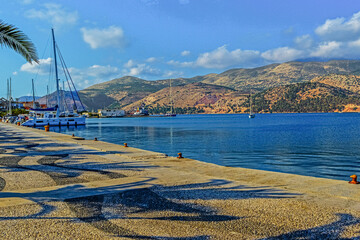 Afternoon view of Argostoli harbor in Kefalonia, Greece, with sailboats, calm blue sea, mountain backdrop and palm tree shadows on the promenade.
