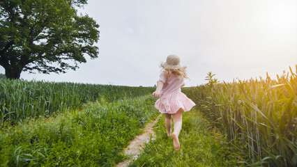 Young girl with blond hair and pink dress running barefoot through a green field at sunset,...