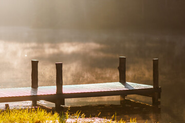 wooden pier extends into a tranquil lake shrouded in soft morning mist. Golden light gently touches the calm water, creating a peaceful and serene natural landscape.

