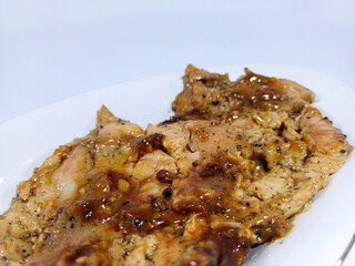 Chicken teriyaki, coated in a rich dark sauce with visible specks of black pepper, shows signs of being grilled and rests on a white plate against a blurred white background. Represents savory chicken