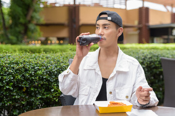 Non binary gay man eating fried chicken outdoors during summer and drinking cola soft drink