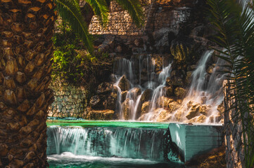 Scenic Waterfall with Flowing Water, Palm Tree Shadows and Stone Wall in Loutraki, Corinth, Greece...