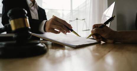 Two hands, one belonging to a legal professional, point to a document for a client to sign, with a gavel and scales of justice nearby.