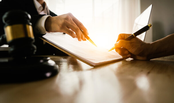 Two hands, one belonging to a legal professional, point to a document for a client to sign, with a gavel and scales of justice nearby. - Powered by Adobe