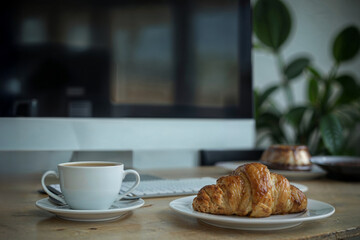 Breakfast on a workspace, a buttery croissant, a cup of coffee, and a small cake, with office equipment and a blurry plant in the background.