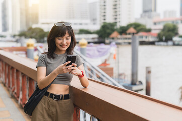 Happy young southeast asian woman teenager blogger world travel explorer using smartphone searching tourism guide. Asia local cultural experience concept at chao phraya river bangkok