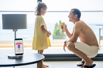 Smartphone with high UV alert in focus, while father in background prepares to apply sunscreen to...