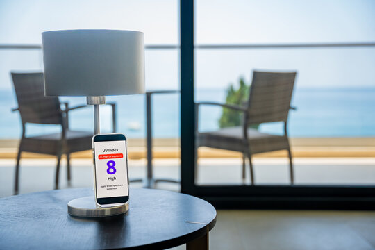 Smartphone displays high UV alert with sunny beach and sea in background. Sun safety reminder during seaside vacation