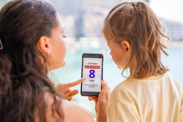 Mother holds smartphone with high UV alert while teaching daughter about sun safety. Educational...