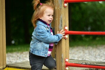 Red hair toddler girl playing in children playground in city park in summer.