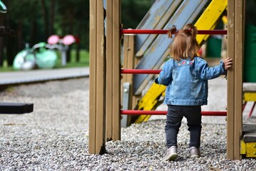 Red hair toddler girl playing in children playground in city park in summer. Little girl standing...