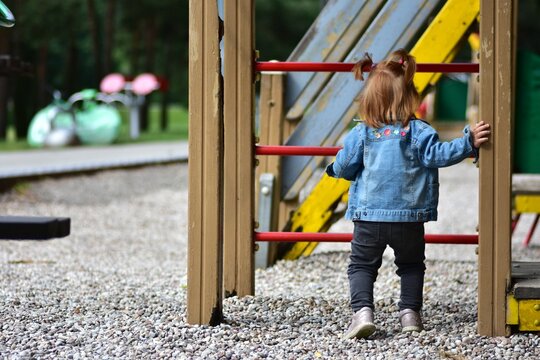 Red hair toddler girl playing in children playground in city park in summer. Little girl standing backwards no face anonymous.