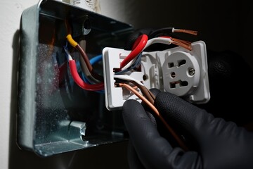 Electrician wearing black gloves connects wires to a white electrical outlet, demonstrating proper installation techniques in a well-lit indoor environment with visible tools and materials present