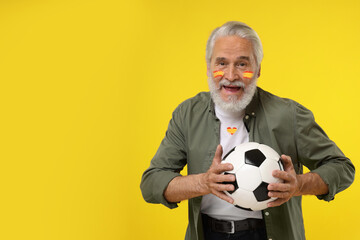 Fan with colors matching Spain flag on his face and t-shirt holding soccer ball against yellow...