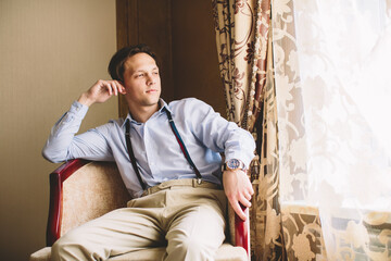 Young Caucasian man in classic outfit relaxing in armchair by window, expressing confidence, emotional depth, self-awareness, timeless style and peaceful modern masculinity.
