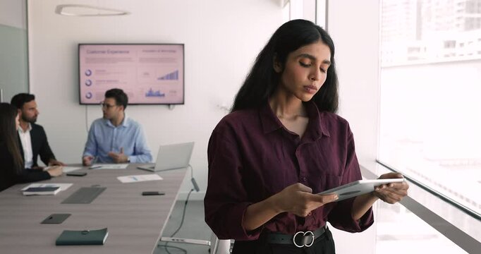 Serious young Indian product manager using tablet in meeting room for online communication, working on tasks, browsing Internet content for analysis. Team of colleagues talking in blurred background