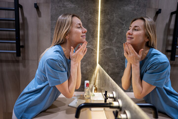 Confident middle-aged woman looks to reflection in bathroom mirror touching face with smile. Natural graceful beauty aging, daily self-care routine, acceptance to wrinkles, skin changes with self-love
