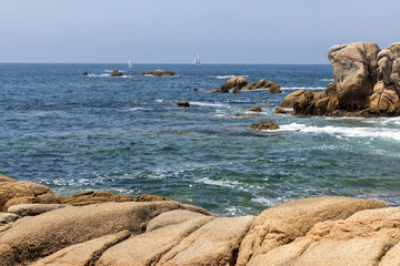 A sailboat is sailing in the ocean near some rocks. The sky is clear and the water is calm