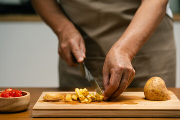 Close-up of Asian man's hands slicing potatoes on a wooden cutting board in the kitchen, preparing...