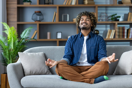 A man meditates peacefully at home, sitting comfortably on a sofa with his eyes closed and a calm expression.