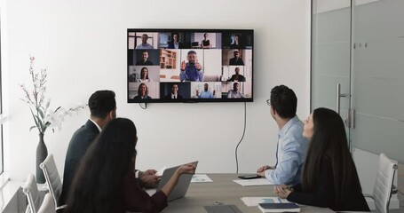 Multiethnic team of colleagues watching online conference on large TV screen, sitting at office table, talking to remote colleagues on group video call, attending corporate webinar on hybrid meeting - Powered by Adobe