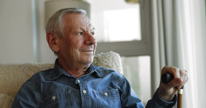Positive thoughtful older senior man with walking disorder using gimp stick, sitting on couch at home, holding knob of walking cane, looking at window away, smiling, thinking on recovery