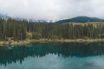 Lake Carezza in Dolomites, Italy, with clear turquoise water, pine forest, and misty mountains in the background.