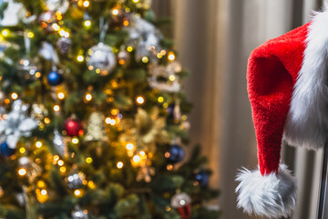 Santa Claus hat hanging on the corner of the TV against the background of a Christmas tree with lit garlands. Blurred background, selective focus.