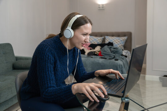 Focused young woman wearing headphones works from home, typing on her laptop and using a mouse, while a cluttered bedroom creates a chaotic backdrop to her everyday life
