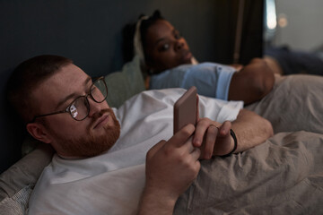 Caucasian young adult man lying in bed using smartphone while Black young adult woman lying beside him looking away, both appearing distant and disengaged, reflecting relationship conflict