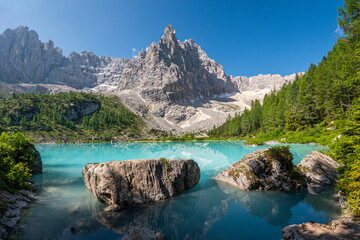 A breathtaking turquoise alpine lake lies beneath a towering rocky mountain peak, surrounded by...