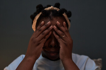 Black woman covering face with both hands, wearing headband, sitting indoors, showing emotional...