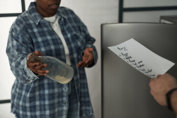 Black woman gesturing with empty glass bottle while standing near refrigerator, Caucasian man...