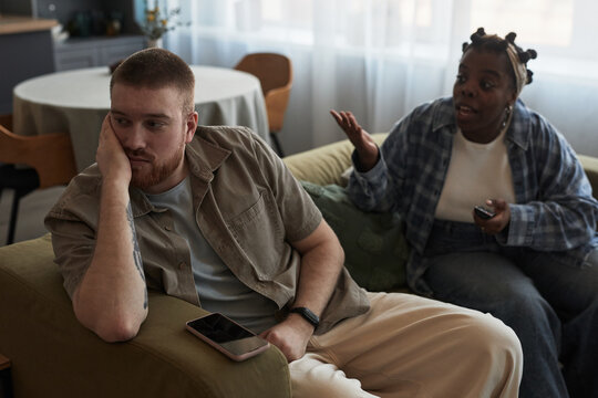 Caucasian young adult man sitting on sofa looking away with hand on face, while Black young adult woman gesturing and talking beside him during conflict in living room