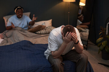Middle aged Caucasian man sitting on bed holding head in hands while middle aged Black woman sitting behind gesturing emotionally, both appearing involved in conflict in bedroom setting
